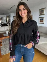 Camisa negra de corte holgado con cuello clásico y cierre frontal de botones. Presenta mangas largas con detalles de paneles laterales y puños en tela escocesa de tonos rosados, blancos y negros.