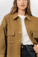 Sobrecamisa de gabardina liviana color crema, con corte oversize, cuello clásico, cierre frontal con botones a presión y dos bolsillos delanteros con solapa.