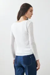 Remera de manga larga, color blanco roto, con cuello bote y textura acanalada. Presenta un efecto de doble capa o transparencia sutil en el escote.