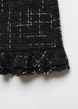 Minifalda negra de tweed con diseño a cuadros blancos, bajo asimétrico con volantes y cierre lateral con cremallera invisible.