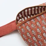 Riñonera de diseño acolchado con estampado de pequeñas hojas blancas sobre fondo color terracota. Cuenta con cierre metálico dorado y correa ajustable del mismo tono.