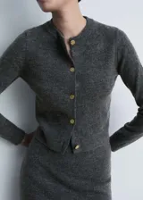 Cardigan de punto gris con cuello redondo y manga larga. Presenta un cierre frontal con botones dorados con textura efecto joya y un diseño de corte recto.