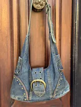 Cartera bandolera de denim azul con apliques de tachas doradas y hebilla metálica.