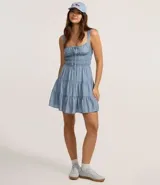 Vestido corto de silueta evasé color celeste, con escote recto y detalle de lazo en el centro. Posee breteles medianos, cintura elástica fruncida y falda con volados escalonados.