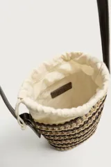 Bandolera vertical con acabado trenzado en tonos tostados y oscuros, cuenta con bolsa interior de tela con cierre de cordón ajustable y correa fina para el hombro.