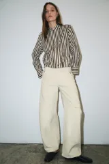 Camisa tipo bomber confeccionada en algodón, con diseño de rayas verticales en tonos marrón y blanco. Presenta cuello perkins, manga larga con puños, bolsillo de plastrón con solapa en el frente, bajo elástico y cierre frontal con botones.
