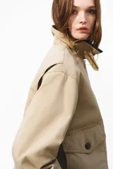 Campera levemente acolchada en lona de algodón gruesa color beige, con cuello y puños vueltos de efecto cuero marrón oscuro. Presenta cierre frontal con botones, solapa rompevientos, bolsillos delanteros de parche con solapa y bolsillos calientamanos.