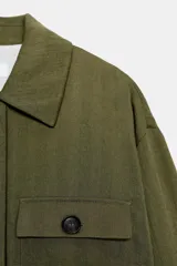 Chaqueta corta de color verde militar, con cuello solapa, bolsillos delanteros con solapa y cierre frontal de botones ocultos. Presenta mangas amplias con vuelta en los puños que muestran un forro a rayas.
