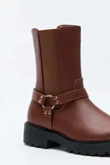 Bota alta color negro, con suela track y taco cuadrado. Presenta una tira con tachas y una argolla metálica decorativa alrededor del tobillo, además de inserciones elásticas laterales.