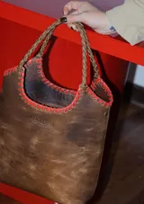 Bolso tipo matera de cuero con textura de reptil, color tostado, con manijas trenzadas y detalles de costura artesanal en los bordes.