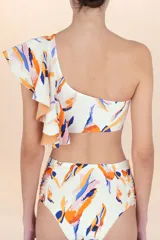 Top de bikini asimétrico, color blanco hueso, con estampado abstracto en tonos naranja, azul y rosado. Presenta un volado amplio en el hombro derecho y tirantes finos ajustables.
