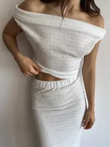 Conjunto de dos piezas en color blanco, compuesto por un top con escote bote y hombros descubiertos, y una falda midi con tajo lateral. Ambas prendas están confeccionadas en tela bambula elastizada con textura arrugada.