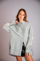 Camisa beige de corte amplio, con cuello clásico, cierre frontal con botones, mangas largas con puños abotonados y bordado floral en las mangas.