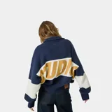 Cardigan de punto grueso color azul marino con cierre frontal de cremallera metálica, cuello tipo polo y corte cuadrado. Presenta un diseño de bloques de color con una franja blanca horizontal en el cuerpo y las mangas, y el logotipo gráfico 'EME' en tono mostaza en el pecho.