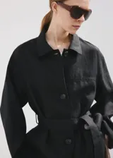 Chaqueta de lino color negro con diseño recto, cuello camisero, manga larga, cierre frontal de botones y cinturón ajustable del mismo material. Incluye forro interior.