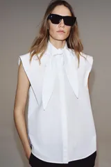 Camisa sin mangas confeccionada en algodón, con cuello redondo y detalle de lazada frontal. Presenta cierre frontal con botones ocultos por solapa y bajo redondeado.