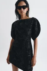 Vestido corto de cuello barco y manga corta abullonada, confeccionado en tejido negro con bordados florales y aplicaciones de abalorios y brillos a tono. Presenta un diseño con volumen en las mangas y cierre de cremallera oculta en la espalda.