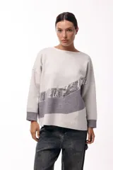 Sweater de punto con calce relajado y mangas amplias. Presenta un diseño de bloques de color en tonos beige y gris, con una franja diagonal de textura metalizada plateada que cruza el frente. Posee puños acanalados en color gris.