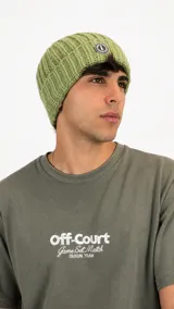 Gorro tipo beanie de tejido de punto acanalado en color verde oliva, con parche circular decorativo en el frente.