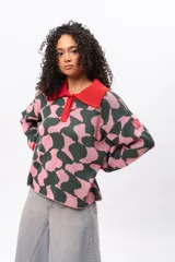 Buzo de tejido de punto con diseño estampado geométrico en tonos verde oscuro y rosado. Presenta cuello tipo polo en color rojo, cierre frontal con botones y puños acanalados en color rojo.