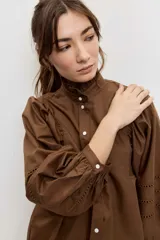 Camisa de manga larga color marrón, con cuello alto con volados, botonadura frontal y detalles calados en las mangas abullonadas.