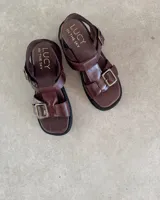 Sandalias de cuero color marrón con suela de goma negra, tiras anchas y hebilla metálica.