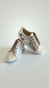 Sneakers de cuero sintético en tonos natural y dorado, con cordones y detalle de aplique artístico en el talón.