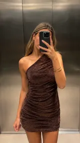 Vestido corto de un solo hombro, confeccionado en tejido de lurex color chocolate con acabado brillante. Presenta un diseño ajustado con frunces laterales que recorren toda la prenda.