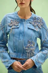 Chaqueta denim de mujer, confeccionada en jean azul claro, con cuello redondo y cierre frontal. Destaca por apliques bordados florales con pedrería y flecos de cristales en la parte frontal, además de detalles de brillos y costuras decorativas a lo largo de las mangas.