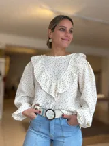 Blusa de tela liviana color crema con estampado floral pequeño en tonos rojizos. Presenta un diseño con volados en el frente y hombros, mangas largas abullonadas con puños elásticos y cuello redondo.