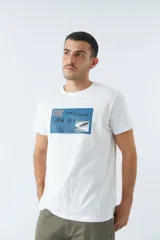 Remera blanca de algodón con cuello redondo y mangas cortas. Estampado frontal con diseño rectangular en tonos azules que incluye texto, coordenadas y la imagen de un avión.