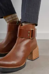 Bota corta de cuero negro con acabado combinado mate y charol. Presenta una tira decorativa con tachas metálicas y hebilla en el tobillo, tacón cuadrado robusto y suela con plataforma.