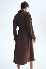 Vestido midi marrón de estilo camisero, con cuello, cierre frontal de botones, hombros caídos y mangas largas. Incluye un cinturón de tela con hebilla metálica de anillas en D.
