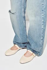Zapato plano tipo bailarina confeccionado en piel de color crudo, con tira en el empeine y hebilla metálica dorada ajustable. Presenta puntera redonda y suela plana.