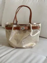 Cartera tipo mini bag de cuero con acabado metalizado dorado, asas de mano y detalle de tira marrón con cierre metálico frontal.