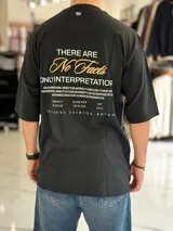 Remera oversize color beige, con mangas cortas y cuello redondo. Presenta un estampado tipográfico en la espalda con la frase "There are no facts, only interpretations" y otros detalles de diseño.