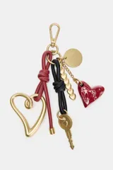 Llavero accesorio con clip metálico, que incluye una llave dorada, un dije de corazón grande de metal dorado, un dije de corazón pequeño esmaltado rojo, un dije redondo dorado, una cadena con dijes de corazón pequeños dorados, y tiras de cuero anudadas en rojo y negro.