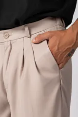 Pantalón sastrero de corte baggy, color beige, con pinzas frontales, cierre de botón y cremallera, y bolsillos laterales.
