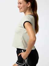 Remera de manga corta estilo crop, color gris claro con detalles en negro en el cuello y en los bordes de las mangas. Presenta un pequeño logo bordado en el pecho.