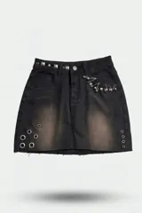 Pollera de jean negro con efecto de lavado localizado, terminación desflecada en el ruedo, tachas cuadradas en la cintura, apliques de alfileres de gancho en el bolsillo y ojalillos metálicos decorativos en la parte inferior.