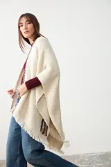 Ruana de tejido de punto en color crudo, con diseño abierto y terminaciones con flecos en el ruedo. Presenta un detalle de trenza decorativa en los bordes frontales y una etiqueta de cuero sintético en la parte inferior.