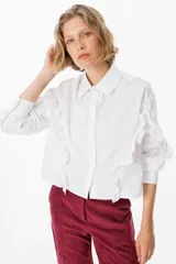 Camisa blanca de corte clásico con cuello camisero y cierre frontal de botones. Presenta volados decorativos que recorren la parte frontal y las mangas, aportando un toque femenino y distintivo.