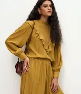 Blusa de manga larga color mostaza con cuello alto y detalle de panel delantero texturizado. Presenta volados aplicados en el busto y puños abotonados.