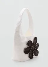 Bolso tipo shopper de fibra natural en color blanco, con diseño de maxi flor tejida en color marrón oscuro en el frente y asa de hombro integrada.
