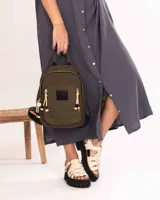 Mochila de nylon color verde oliva, con compartimento principal y bolsillo frontal con cierre metálico. Presenta detalles de cordones decorativos en los tiradores de los cierres, una grifa de goma con el logo de la marca y correas ajustables.