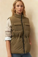 Chaleco puffer color crema, con cuello alto, cierre frontal y cuatro bolsillos con solapa. Cuenta con cintura ajustable con cordón.