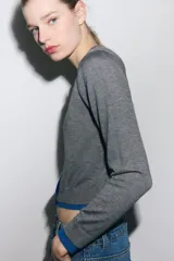 Chaqueta de punto gris de manga larga, cuello redondo y cierre frontal con botones oscuros. Presenta ribetes y detalles en color azul eléctrico en el frente, puños y ruedo.