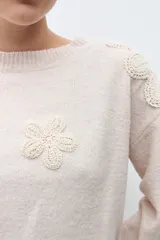 Sweater de punto en color beige melange, con cuello redondo y mangas largas. Presenta apliques de flores tejidas en relieve sobre los hombros y el pecho.
