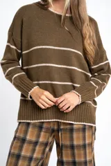 Sweater de lana con diseño de rayas horizontales blancas sobre fondo verde oliva. Presenta cuello redondo, mangas largas con puños acanalados y un corte oversize con tajos laterales en el ruedo.