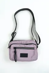Bandolera de nylon color lila con múltiples compartimentos con cierre, tira ajustable negra y detalle de etiqueta engomada en el frente.